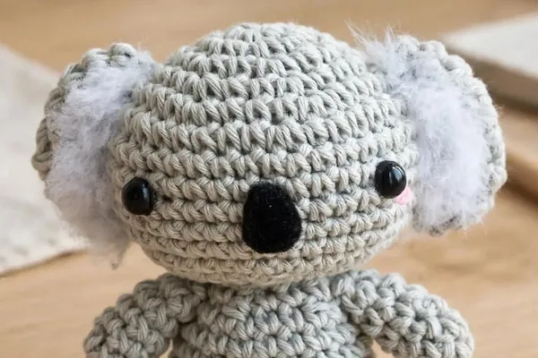 A small gray crocheted koala with fluffy white ears, a black felt nose and pink cheeks, sitting on a wooden table