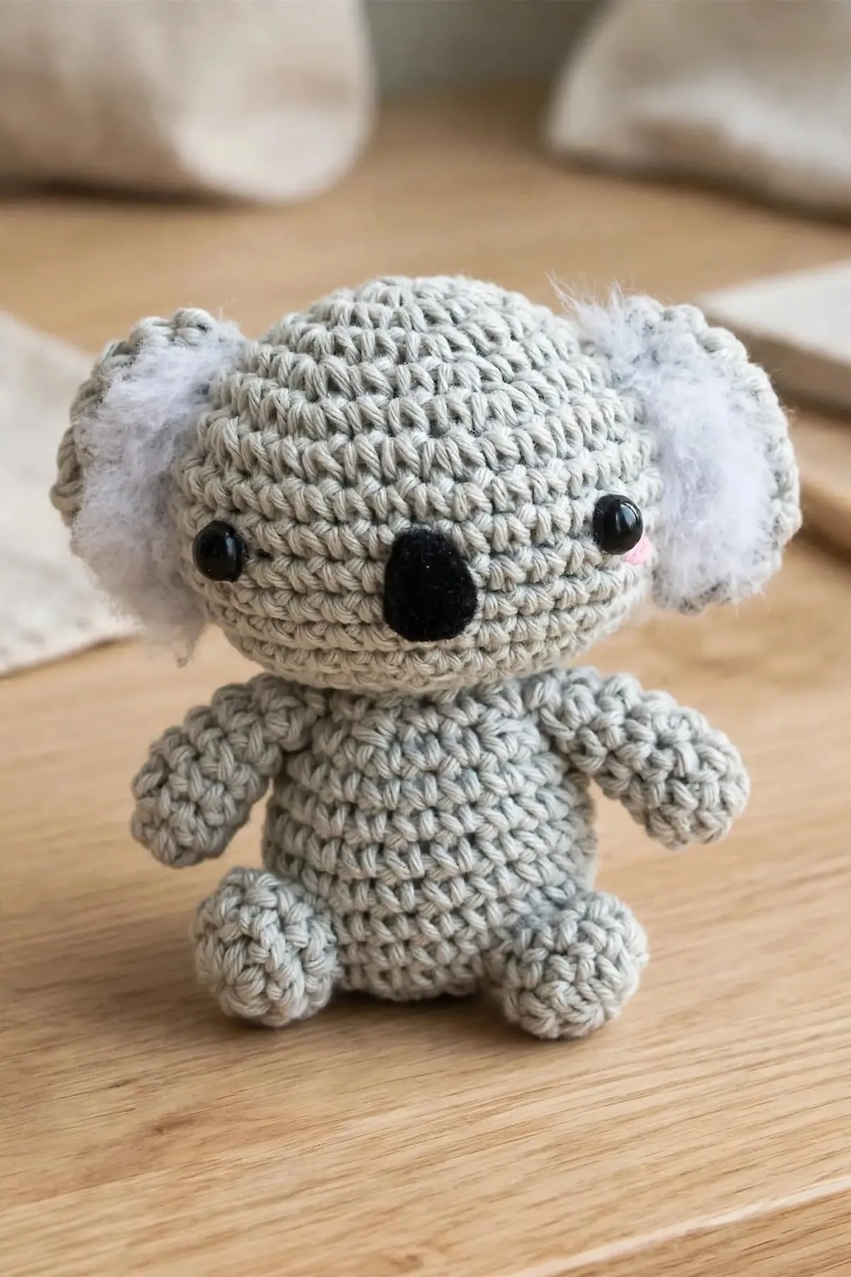 A small gray crocheted koala with fluffy white ears, a black felt nose and pink cheeks, sitting on a wooden table