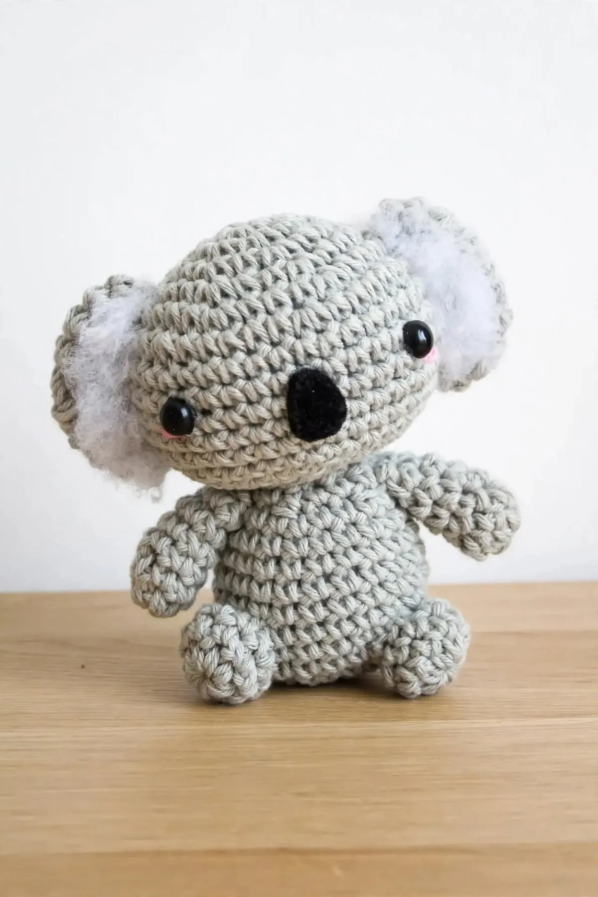 The crocheted mini koala sitting against a white wall, with its arms out and fluffy white ears