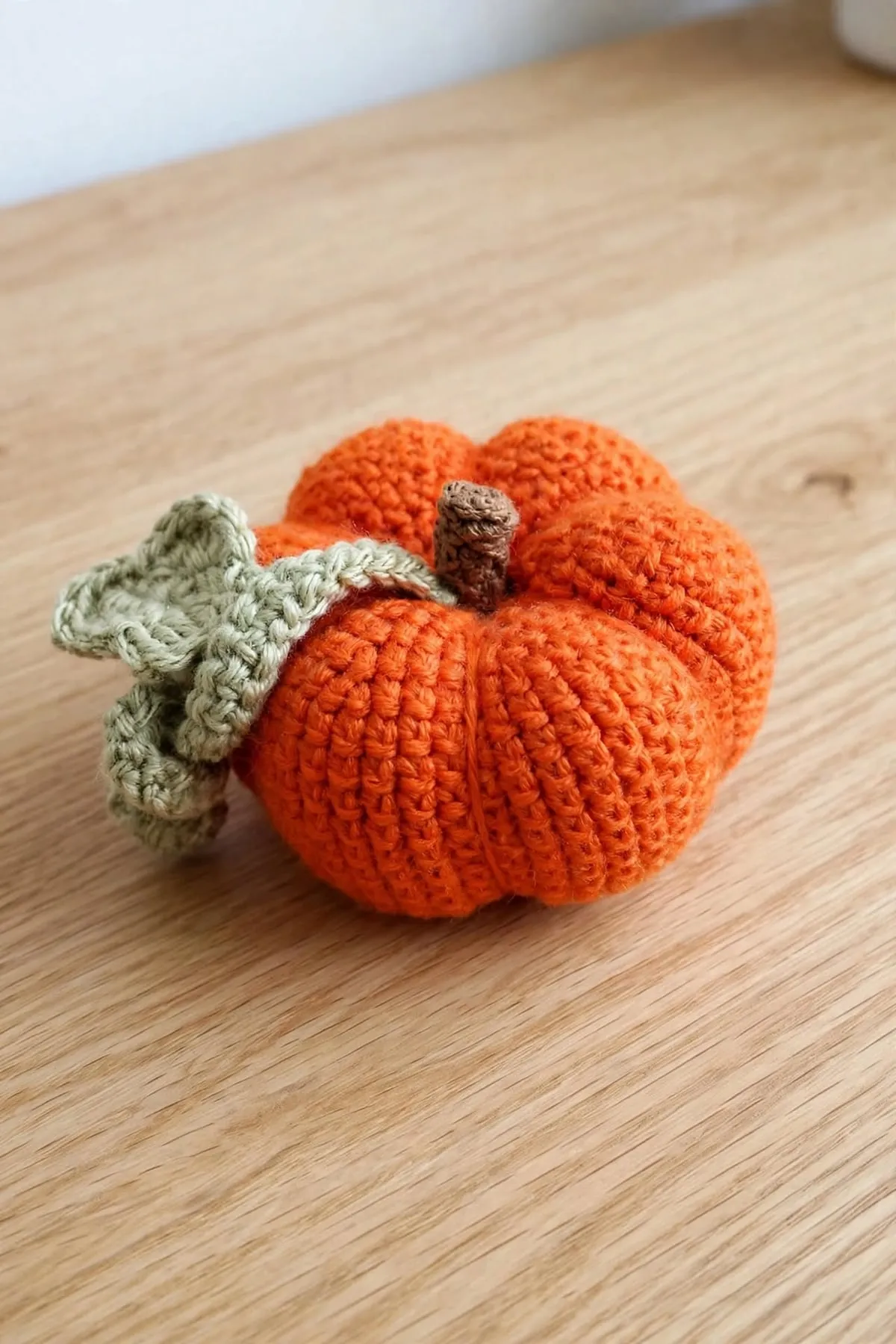 A crocheted pumpkin with a sage green leaf draped over the top beside its brown stem