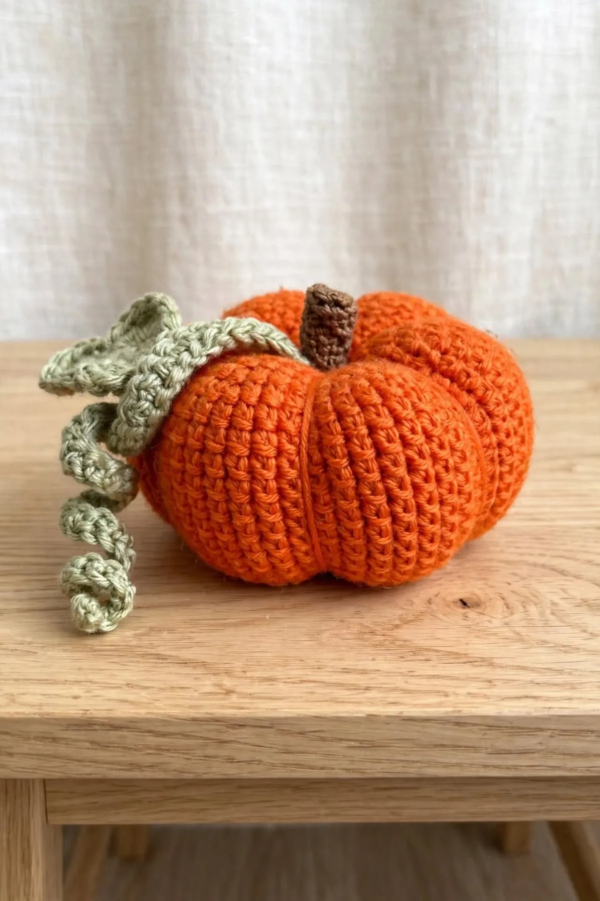 A crocheted pumpkin with a curly green vine hanging over the edge of a wooden stool